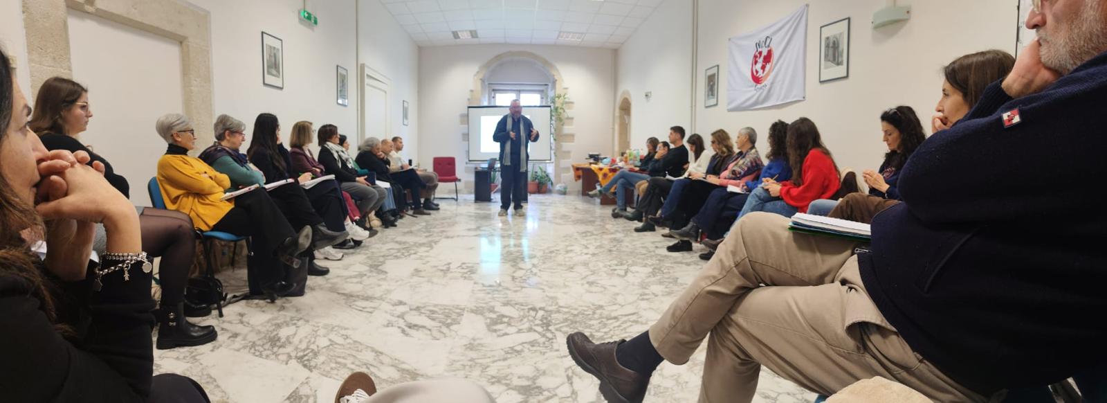 Persone disposte sui due lati di una sala durante un incontro di ascolto e partecipazione